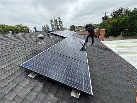 A technician rinsing a long row of solar panels on a shingle roof with a water-fed pole